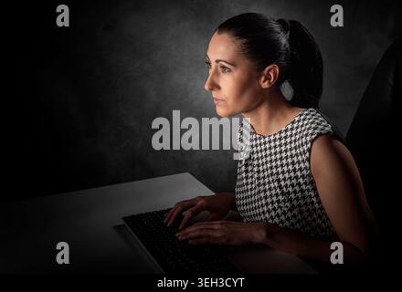 A professional woman sits at her desk in a dark office, focused on her computer screen. Stock Photo