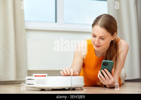 A woman sits on a chair in a room and turns on a robot vacuum cleaner using a smartphone Stock Photo