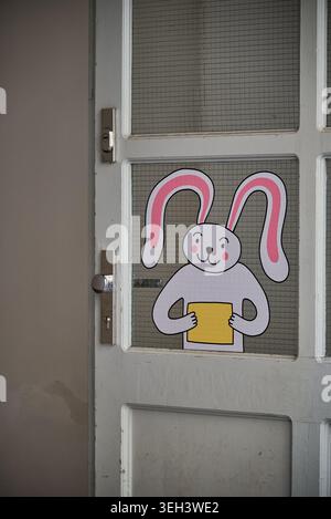 Cute cartoon bunny with pink ears holding a yellow card on a glass door window, cozy home entry, simple festive decoration concept. Stock Photo