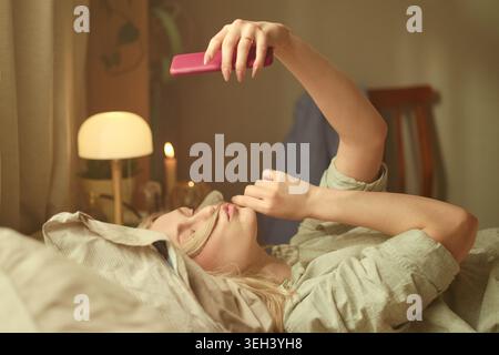 Caucasian young adult woman lying in bed and using smartphone, scrolling late at night with insomnia concept. Suitable for sleep disorder, screen addiction, digital lifestyle campaigns Stock Photo