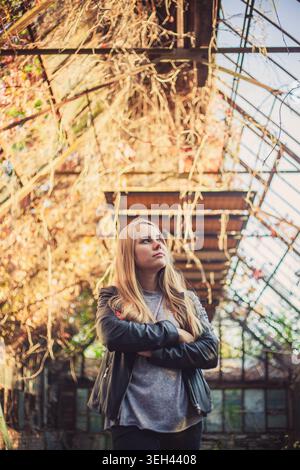 Pensive young woman with crossed arms looks up inside old greenhouse. Warm golden autumn light creates thoughtful mood. Low angle shot with soft focus Stock Photo
