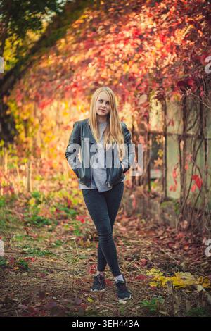 Young blonde woman poses confidently in autumn park. She wears black leather jacket and dark pants. Colorful red and yellow foliage creates beautiful Stock Photo