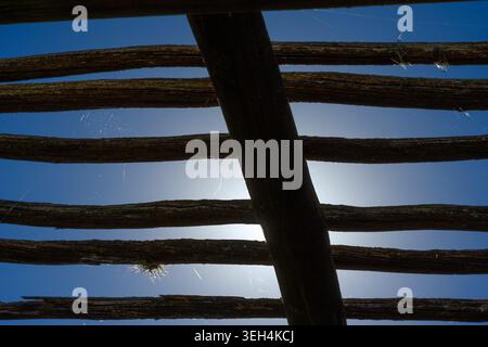Sunlight filters through weathered wooden beams. Blue sky contrasts with rough, aged wood structure. Webbed threads catch light between dark slats. Stock Photo