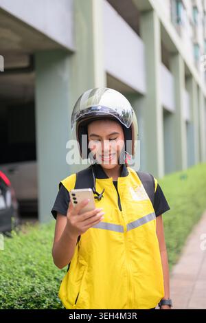 A vertical medium shot captures a cheerful young Isan Thai female delivery courier looking down at her smartphone screen with a warm smile while stand Stock Photo
