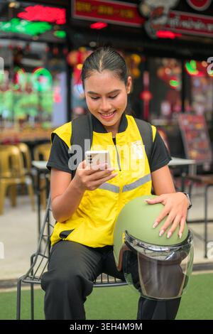 A vertical medium shot captures a happy young Isan Thai female delivery courier sitting down on an outdoor chair while checking her smartphone with a Stock Photo