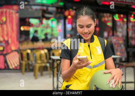 A horizontal medium shot captures a happy young Isan Thai female delivery courier sitting down on an outdoor chair while checking her smartphone with Stock Photo