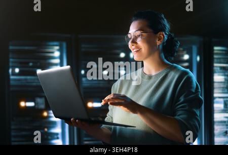 A confident young woman IT professional in glasses stands in a high-tech server room, smiling while working on her laptop. The dim, atmospheric lighti Stock Photo