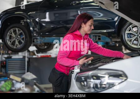 Female car mechanic inspects a vehicle while working in an auto repair shop. She uses diagnostic tablet and equipment for maintenance tasks. Stock Photo