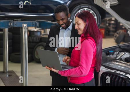 Woman mechanic standing next to an open-hood car, explaining the vehicle maintenance checklist to a client using a laptop. Stock Photo