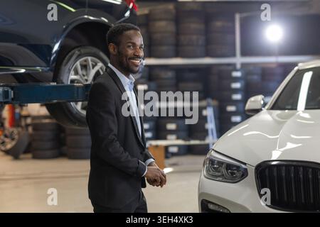 A professional man stands in an auto repair shop surrounded by cars and mechanics working. Stock Photo