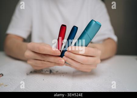 Closeup of hands holding three different colored heat-not-burn electronic cigarettes above table with used cleaning sticks and residue, illustrating Stock Photo