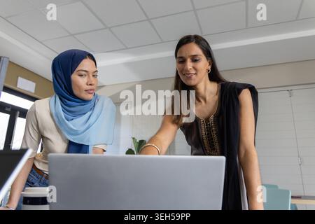 Diverse female colleagues in headscarf and embroidered tunic collaborating on laptop at table Stock Photo