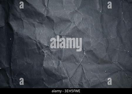 Crumpled black paper texture in close-up, showing dark wrinkles, creases, rough folds and small dust marks as a grunge abstract background. Stock Photo