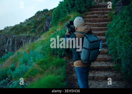 A behind-the-scenes shot from a back perspective shows a male content creator or filmmaker shooting video in a natural mountain environment. Wearing a Stock Photo