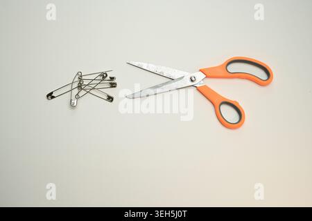Orange-handled scissors with metal safety pins arranged on a plain background, a minimalist still life of sewing and household fastening tools. Stock Photo