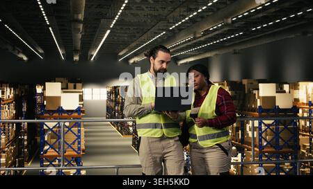 Two people working together on logistics in industrial warehouse, supply chain management. Wearing safety vests, handle packages and plan delivery operations using tracking system on laptop. Stock Photo