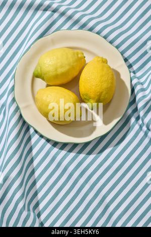 three vibrant yellow lemons placed on a minimalist white ceramic plate. The plate rests on a textured, wrinkled blue and white striped linen tableclot Stock Photo