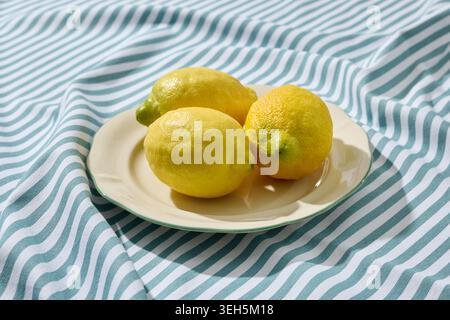 three vibrant yellow lemons placed on a minimalist white ceramic plate. The plate rests on a textured, wrinkled blue and white striped linen tableclot Stock Photo