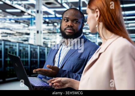 Data center system administrators doing brainstorming, reviewing network configuration, doing maintenance. Server farm coworkers collaborating, scanning hardware logs on laptop, tracking glitches Stock Photo