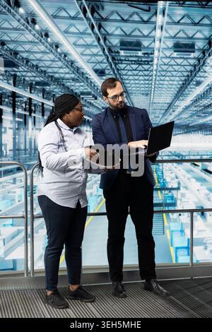 Man and coworker checking silicon wafer hardware defects in industry 4.0 manufacturing plant with laptop. Managers assessing smart factory semiconductor substrate yields using notebook Stock Photo