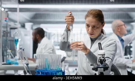 Assay development specialist in lab pipetting blue liquid into test tube to analyze it. Molecular biologist transferring chemical sample to prepare for immediate spectroscopic analysis, camera B Stock Photo