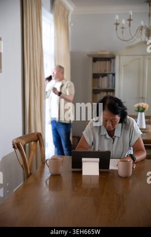 Senior Asian Chinese couple sitting at dining table, woman using tablet, man talking on smartphone Stock Photo