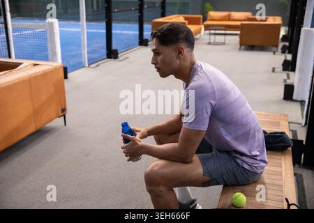 Adult man sitting on bench beside court in light purple shirt holding blue bottle, tennis ball Stock Photo