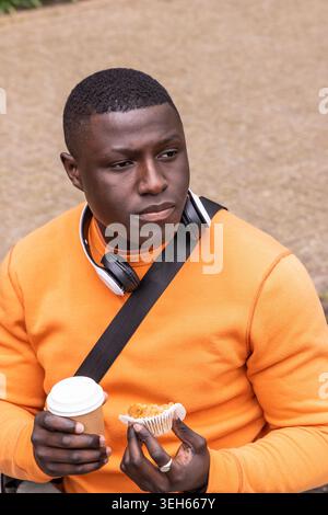 African American man sitting along path wearing orange with headphones holding coffee and muffin Stock Photo