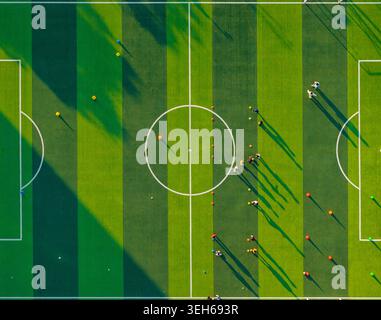Aerial view captures players training on a bright green artificial turf soccer field under a late afternoon sun casting long shadows Stock Photo
