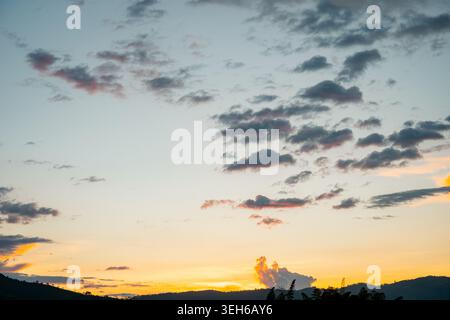 Golden hour light illuminating scattered clouds above dark mountain silhouettes, creating a moody and tranquil evening sky with hints of orange and bl Stock Photo