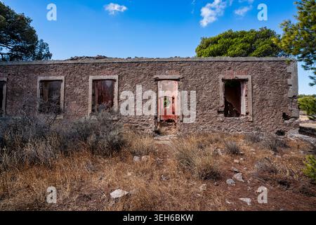 Front view of a ruined stone mining-era building in Lavreotiki, Greece, surrounded by dry vegetation and pine trees under a bright Mediterranean sky. Stock Photo