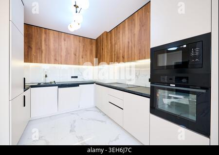 Stylish modern kitchenette with integrated oven tower, induction cooktop and sink set into corner workspace. Contemporary residential interior with dark backsplash and refined materials Stock Photo
