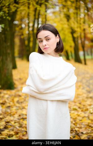 Portrait of young woman posing in white knit sweater in autumn park, standing among golden leaves and trees. Ideal for fashion, cozy lifestyle, beauty, seasonal campaigns, and advertising use Stock Photo