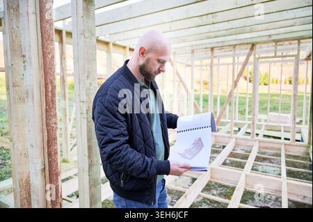 Builder studies architectural plans inside timber house frame, reviewing construction details and coordinating layout during early stage of residential project Stock Photo