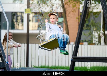 Girl pushing boy on swing at playground, both smiling and playing together outdoors, active kids enjoying fun. Concept of friendship, support, carefree childhood Stock Photo