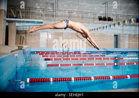 Professional swimmer diving into pool during training, male athlete in streamlined position jumping from starting block in indoor sports facility with lane lines and clear blue water Stock Photo