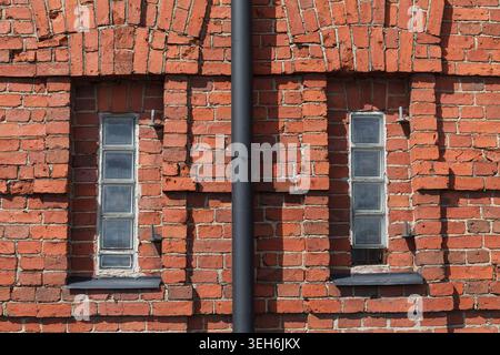 Close-up of weathered red brick wall with two narrow windows and downpipe on old building. Stock Photo