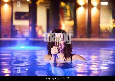 Young woman in pool at night raising champagne glass. Concept of celebration, luxury lifestyle, relaxation and premium spa resort experience with cozy lighting and modern setting Stock Photo