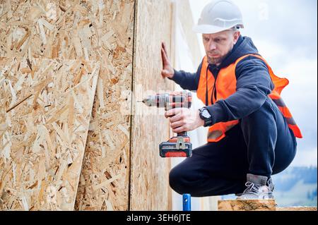 Worker securing wall boards on timber frame while balancing on scaffolding, showing facade installation, site safety and efficient use of modern construction equipment Stock Photo