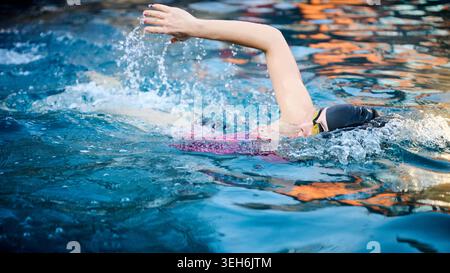 Woman swimming freestyle in pool, side view with splashing water and vivid reflections. Concept of movement, energy and outdoor fitness activity Stock Photo