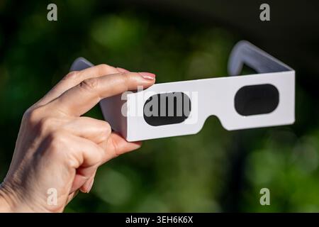A female hand holds a pair of special glasses for observing the sun, against a dark green background Stock Photo