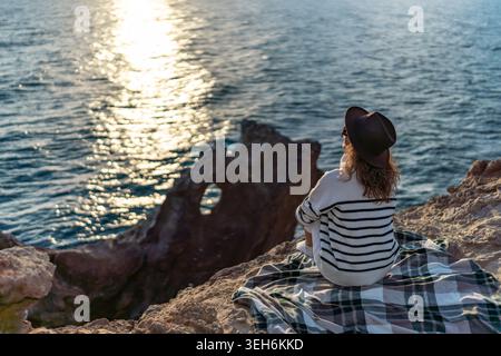 Woman sunset ocean view sitting on plaid blanket on rocky cliff contemplating nature Stock Photo