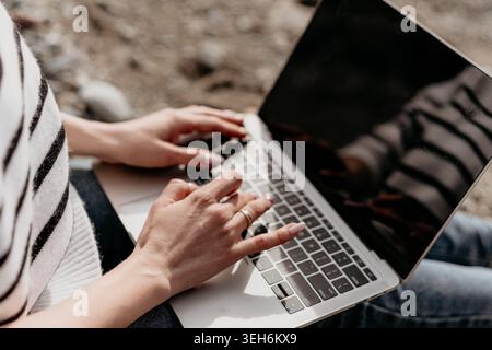 Typing laptop freelance worker sitting on a coastal beach using a portable computer for remote work during a sunny day Stock Photo