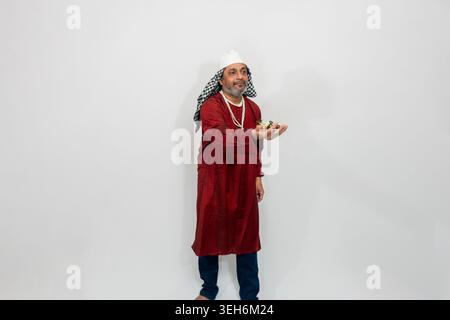 Indian man wearing traditional attire, including a red tunic, white headdress, and patterned scarf, posing against a plain background. Stock Photo