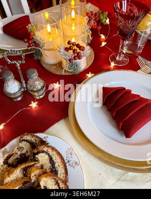 Elegant Christmas table with a tree-folder red napkin, golden charger plate, warm fairy lights, candles, and traditional sweet holiday bread. Stock Photo
