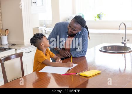 African American man helping schoolboy in orange tee with homework at island using yellow pencil Stock Photo