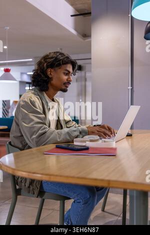 Indian man wearing light green jacket, working on laptop with notebook and phone at coworking table Stock Photo
