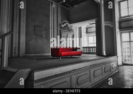 Modern red leather sofa in dark atmospheric abandoned room with dramatic spotlight. Minimalist luxury couch in moody rustic architectural hall. Stock Photo