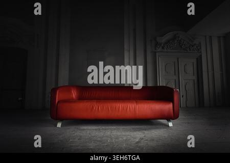 Modern red leather sofa in dark atmospheric abandoned room with dramatic spotlight. Minimalist luxury couch in moody rustic architectural hall. Stock Photo