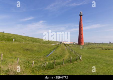 Den Helder, Netherlands. A wide scenic view of the historic red Lange Jaap lighthouse standing against a blue sky, surrounded by green grassy dikes an Stock Photo